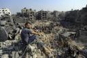 A man sits on the rubble as others wander among debris of buildings that were targeted by Israeli