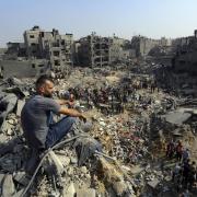 A man sits on the rubble as others wander among debris of buildings that were targeted by Israeli